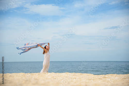 Cheerful asian woman barefoot walking on tropical summer beach. Woman walking along wave of sea water and sand on the beach. Enjoyment barefoot walk outdoor with freedom. Relaxation Travel Concept.