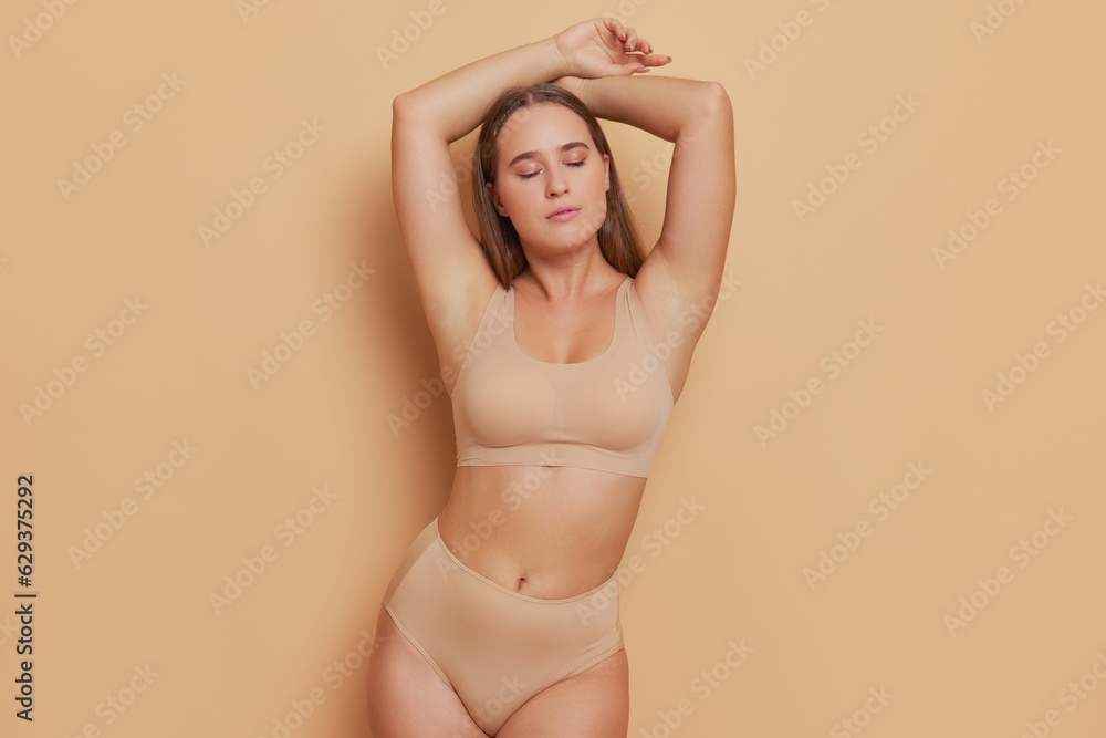 Graceful young woman wearing beige bra and panties stands at light brown background, hands over her head, natural beauty concept, copy space - obrazy, fototapety, plakaty 