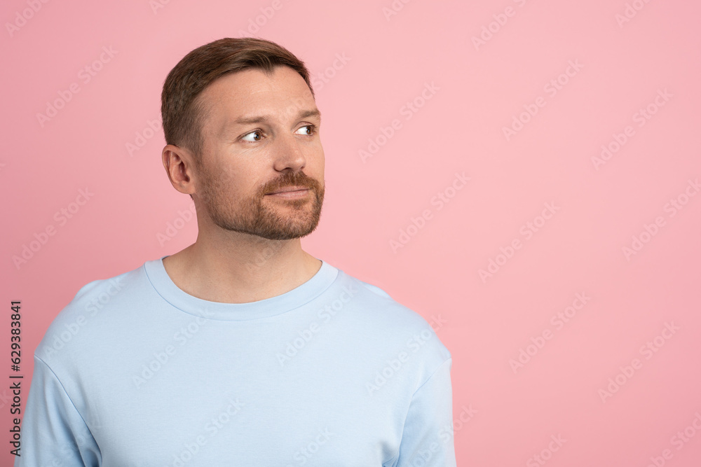 Interested man with perfect skin, well-groomed stubble on face looking away on pink studio background for announcement, advertising, offer, sale. Male wearing blue t-shirt slightly smiling copy space