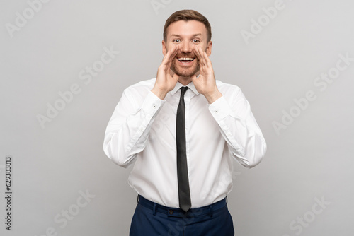 Happy office worker man shouting with hands near mouth gesture on grey background. Advertisement banner, poster. Smiling businessman in white shirt with black tie screaming and looking at camera.