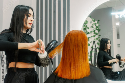 Woman hairdresser stylist of oriental appearance works in beauty salon and makes hairstyle for client. Red hair dye. Dry hair with hairdryer. Looks in mirror. Sits in chair.