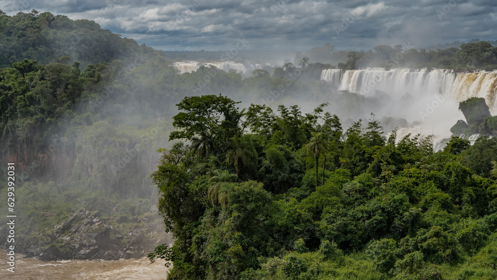 The waterfall flows collapse from the edge of the plateau. Spray, fog. Lush tropical vegetation all around. Clouds in the blue sky. Iguazu Falls. Argentina. The waterfall flows collapse from the edge of the plateau. Spray, fog. Lush tropical vegetation all around. Clouds in the blue sky. Iguazu Falls. Argentina.