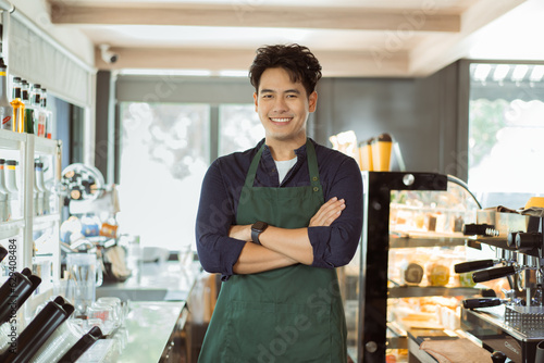 Young Asian male barista in apron having arms folded and smiling while looking at camera with copy space