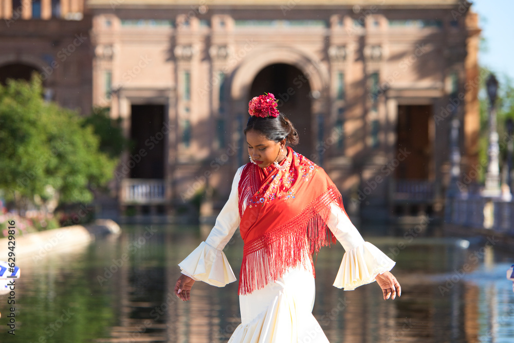 Obraz premium Young black woman dressed as a flamenco gypsy in a famous square in Seville, Spain. She is wearing a beige dress with ruffles and a red shawl and is in front of a canal in the square.