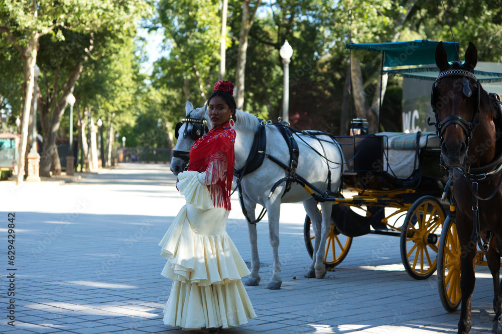 Obraz premium Young black woman dressed as a flamenco gypsy in a famous square in Seville, Spain. She is wearing a beige dress with ruffles and a red shawl and is standing next to a horse-drawn carriage.
