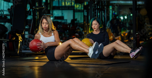 Two Asian women gym partner doing seated oblique twist exercise with weight medicine ball on the floor which strengthen the abs abdominal and core body muscle