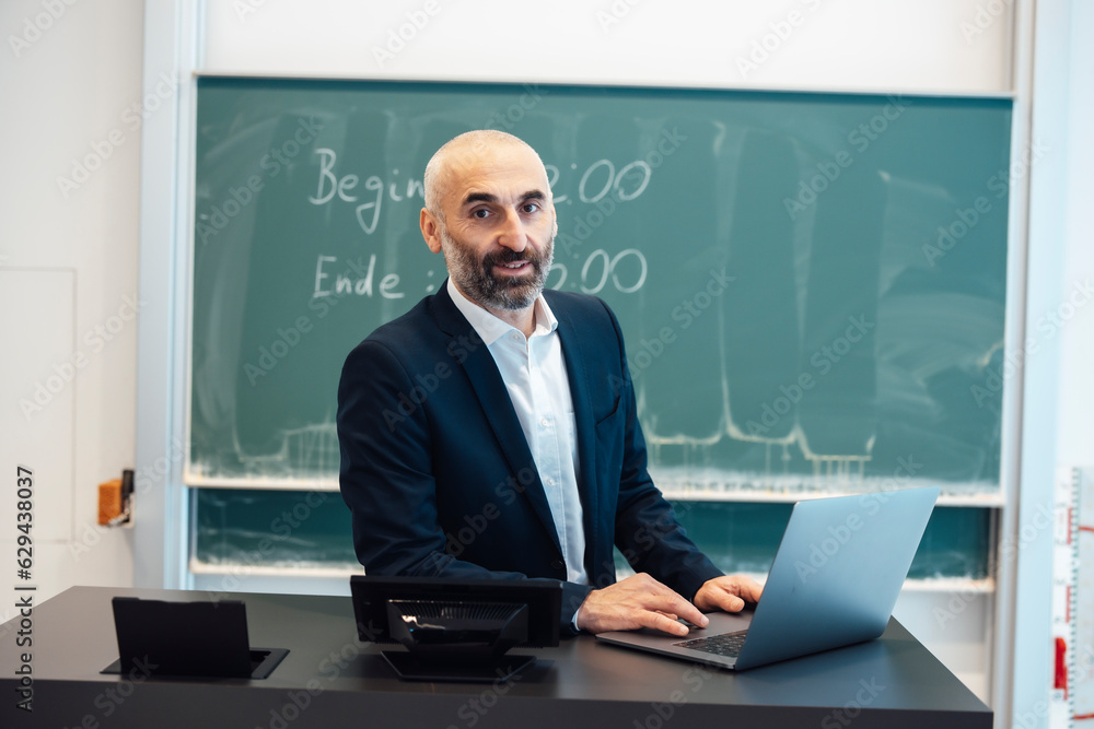 Professor sitting at desk with laptop in lecture hall Professor sitting at desk with laptop in lecture hall