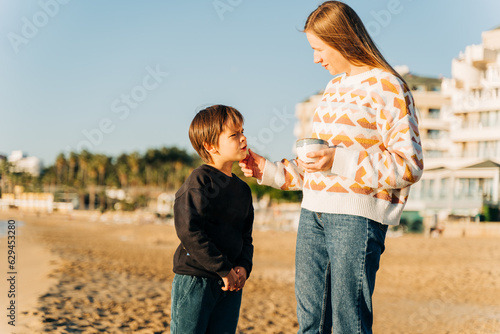 Young beautiful woman mother drinking coffee and spending free time on the winter sandy beach with her school kid son boy. #629453280