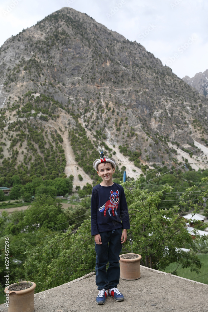 Tourist child, portrait of cute boy, fun kid, toddler in beautiful mountains in Chitral, Pakistan. Tourist child in traditional national folk hat, costume. Fashion boy in mountains. Nature of Pakistan