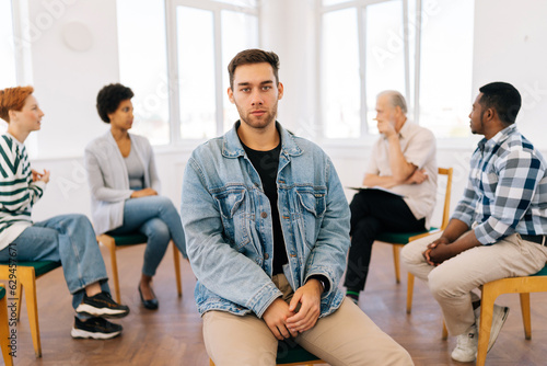 Portrait of sad Caucasian man looking at camera with serious expression while group of multicultural and different ages people sitting in circle, talking in background during therapy session. © dikushin