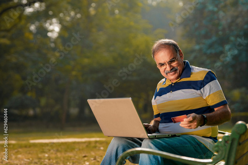 Senior indian man using laptop and bank card at park. older man with technology. © Niks Ads