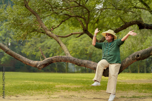 Happy senior indian woman wearing hat and sitting on tree branch at in the green forest nature.