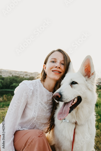 a girl with braces looks at the camera and hugs a white dog against the background of the city at sunset. a woman hugs a white shepherd. © Валерий Георгиян