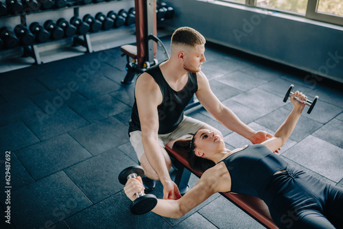 In the gym, a knowledgeable male personal trainer provides guidance to a woman lifting a dumbbell. Fitness Training Male Personal Trainer Helps Woman with Dumbbell Exercise