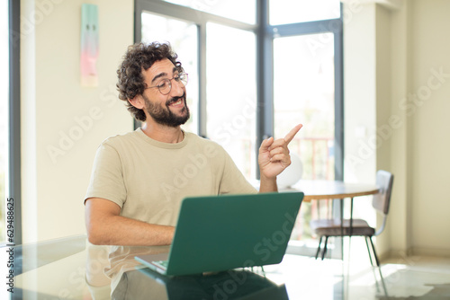 young adult bearded man with a laptop smiling cheerfully, feeling happy and pointing to the side and upwards, showing object in copy space