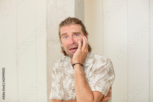 young blond adult man open-mouthed in shock and disbelief, with hand on cheek and arm crossed, feeling stupefied and amazed photo