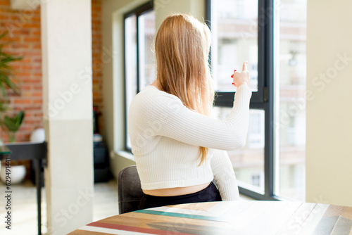 young pretty woman standing and pointing to object on copy space, rear view. home interior