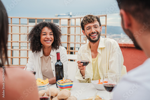 Canvas Print Gathering of people enjoying the good weather sitting at the table, talking, drinking wine at rooftop party