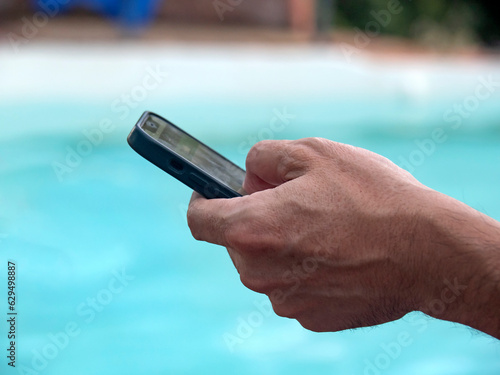 Mano de hombre sujetando un teléfono móvil junto a la piscina / Man's hand holding a mobile phone by the pool