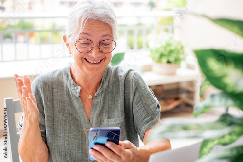 Video call concept. Happy senior woman with eyeglasses having video call on mobile phone sitting outdoor on terrace, elderly lady using modern technology and wireless connection