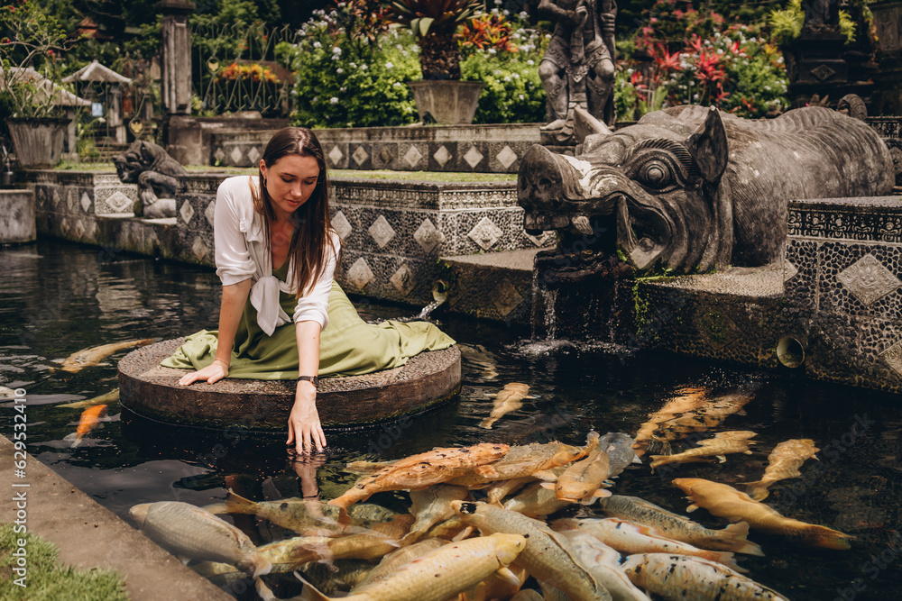 Obraz premium Beautiful girl sitting and admiring pond koi fishes. Smiling girl feeding carps in Ubud Water Palace