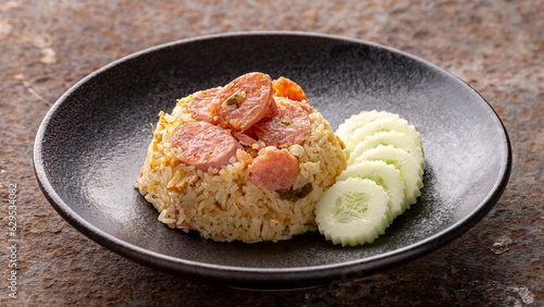 fried rice with fermented pork, sour pork, pickled pork sausage and egg serve with sliced cucumber in black ceramic plate on rusty texture background, Khao Pad Naem, Thai food photo