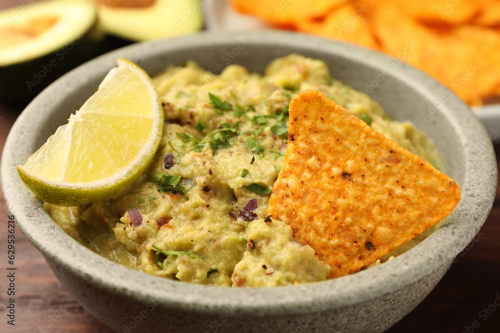 Bowl of delicious guacamole, lime and nachos chips on wooden table, closeup