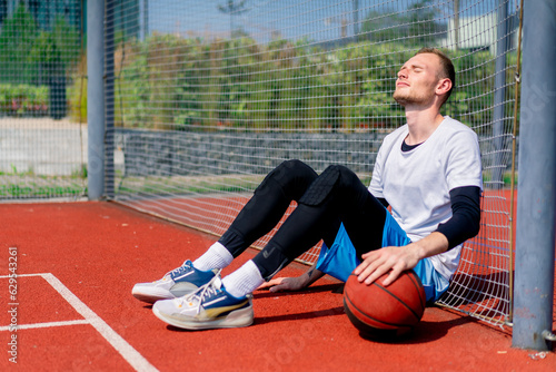 A tall guy basketball player with a ball in his hand sits leaning on the net of a basketball court during practice in the park  © Guys Who Shoot