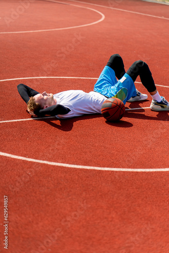Tall guy basketball player lying on a basketball court in the park along with a basketball resting during practice 