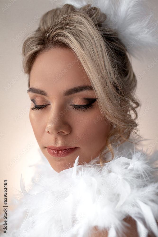 Portrait of a beautiful bride with a wedding hairstyle dressed in a white dress with feathers. A girl with a professional makeup against the wall