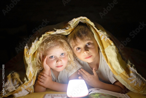 Two blond cute childrem, boy and girl, siblings, lying under the cover in bed, reading book together with small light photo