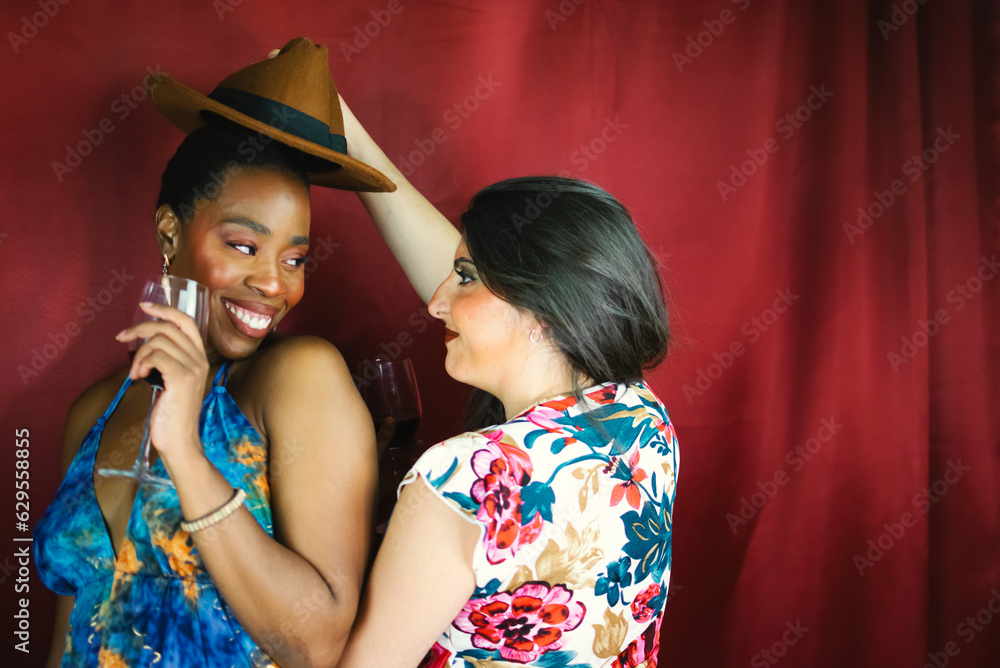 Naklejka premium Two female multiracial friends with hat, smiling, dancing and drinking wine