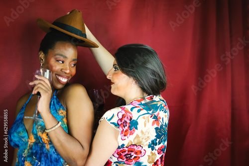Two female multiracial friends with hat, smiling, dancing and drinking wine