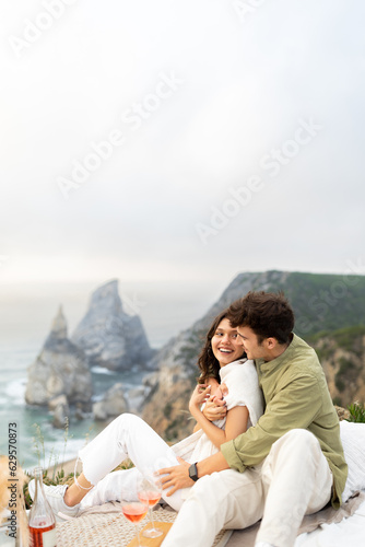 Couple in love having picnic, embracing, drinking wine, enjoying date with beautiful view on ocean shore, vertical shot