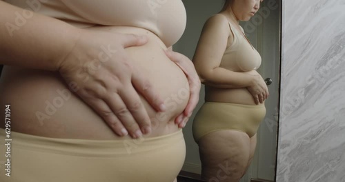 Asian oversize woman in front of a mirror looks at her body. Excess belly fat, Unhealthy lifestyle, Diet concept.
