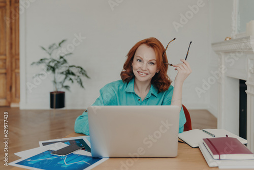 Happy redhead woman in coworking space works on laptop, holds spectacles, browses web, communicates online. Blue shirt, paper documents.