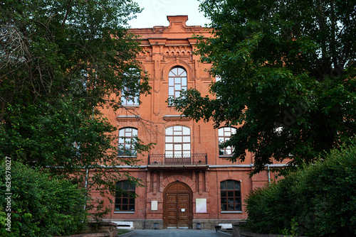 Part of old three storeyed red brick building standing among green trees and bushes on summer day in urban environment photo