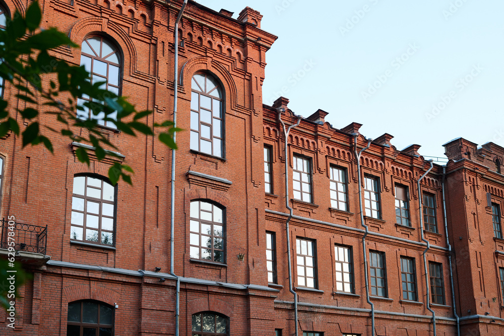 Part of red brick historical building of three storeys standing in front of camera in urban environment with blue sky above