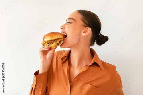 Satisfied young woman biting tasty burger with pleasure, enjoying cheat meal over white wall background