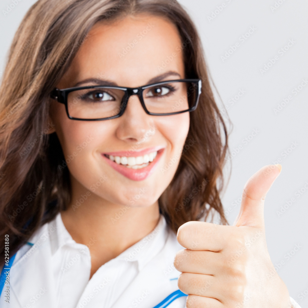 Happy smiling cheerful female doctor with thumbs up gesture, over grey background Happy smiling cheerful female doctor with thumbs up gesture, over grey background