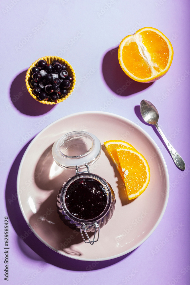 Blackcurrant jam with orange in a glass jar on a lilac background.