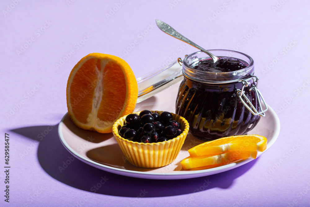 Blackcurrant jam with orange in a glass jar on a lilac background.