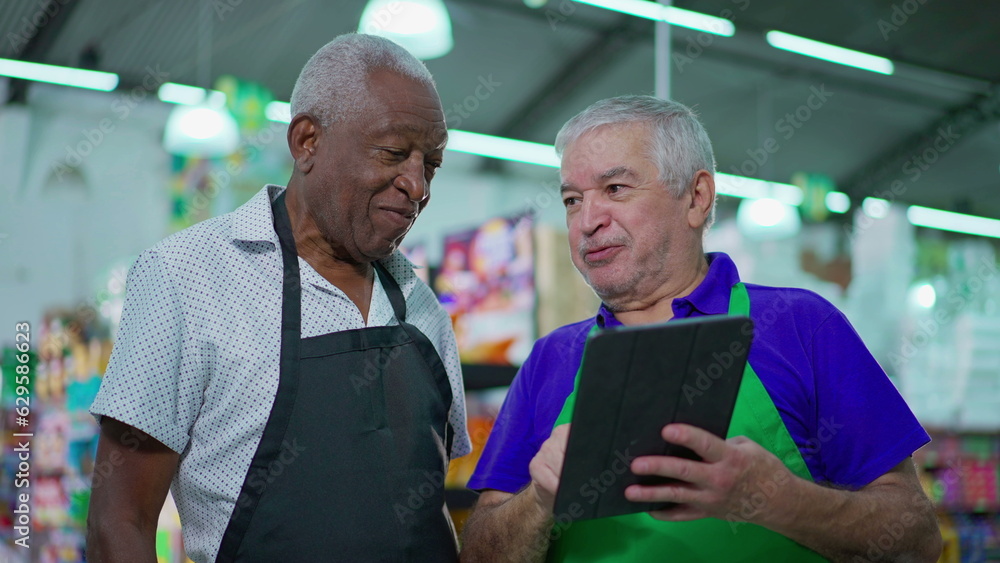 Two happy diverse senior staff of frocery store checking inventory operations with tablet device, older employees of supermarket laughing and smiling at workplace Two happy diverse senior staff of frocery store checking inventory operations with tablet device, older employees of supermarket laughing and smiling at workplace