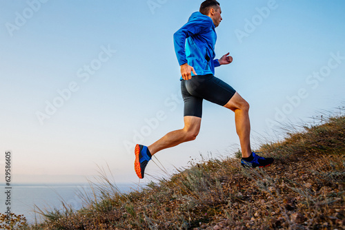 side view male runner athlete running uphill in blue jacket and black tights, background of sky and sea photo