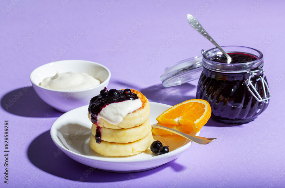 Breakfast of syrniki with sour cream and blackcurrant jam with orange on a lilac background.