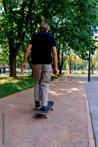 young guy skater rides a skateboard on the path of the city park against the background of trees and sky