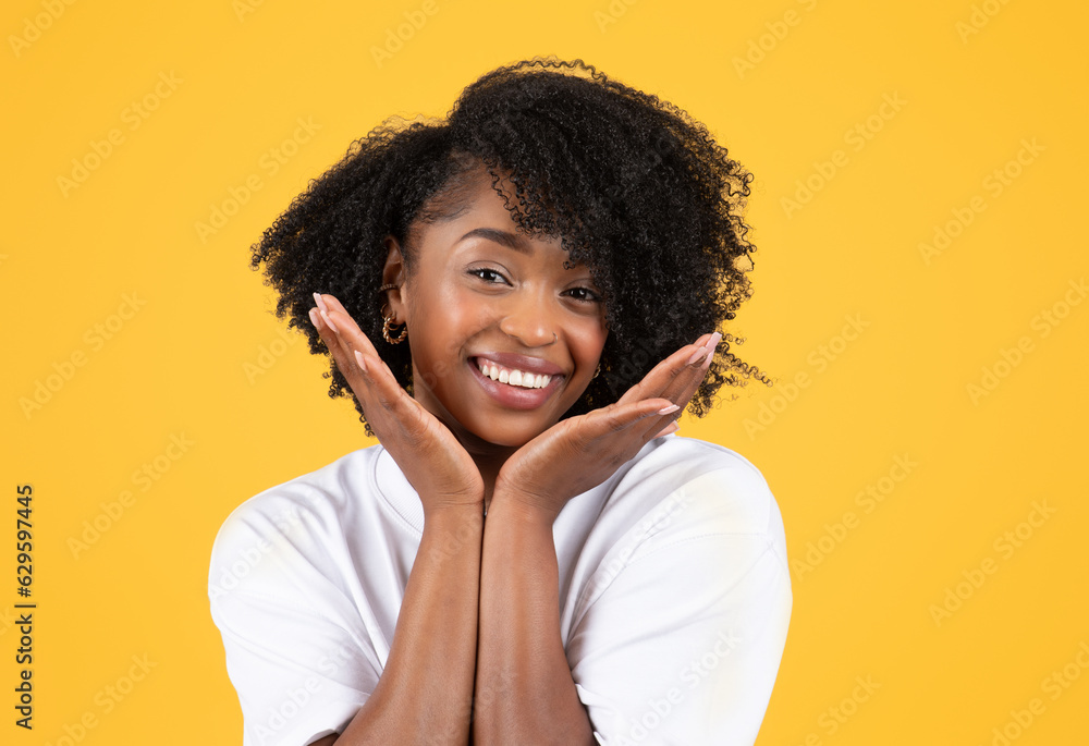 Positive young black curly lady in white t-shirt touches face with hands, enjoys beauty care Positive young black curly lady in white t-shirt touches face with hands, enjoys beauty care