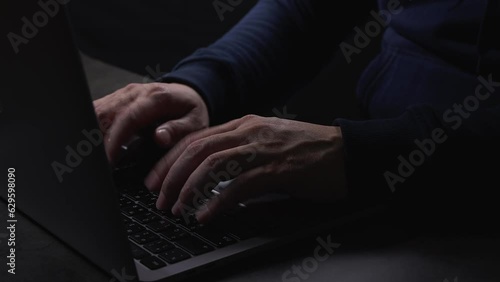 Man wearing hood jacket typing on laptop keyboard (English and Thai language buttons) buttons while working at night via Internet. Online communication and workplace concept.