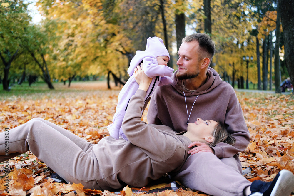 Fototapeta premium Young parents are sitting on a plaid with a newborn baby in an autumn park. A mother raises a child in her arms, a father kisses her daughter