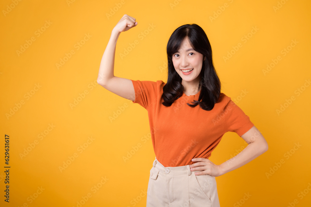 Confident young Asian woman in her 30s raises a fist up hand sign gesture, wearing an orange shirt on yellow background. Empowerment and celebration concept. Empowerment and feminism concept. Confident young Asian woman in her 30s raises a fist up hand sign gesture, wearing an orange shirt on yellow background. Empowerment and celebration concept. Empowerment and feminism concept.